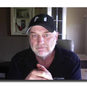 May include: A man wearing a black baseball cap with white accents and a black long-sleeve shirt. The man has a white beard and is looking at the camera. The Columbia logo is visible on the shirt.