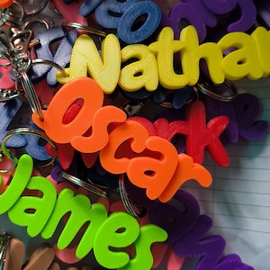 May include: A collection of colorful name keychains in various colors, including orange, yellow, green, and purple. Each keychain features a different name, such as "Oscar" and "James", in a bold, 3D font. The keychains are attached to silver keyrings.