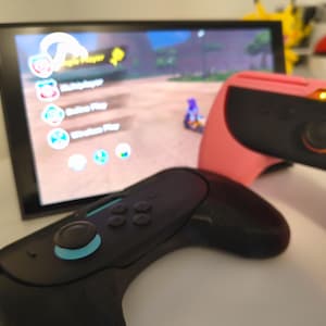 Puede incluir: Una consola de videojuegos negra y rosa con mandos. La pantalla muestra un menú de juego con opciones para un jugador, multijugador, juego en línea y juego inalámbrico. Una figura de Pikachu y una Pokeball son visibles en el fondo.