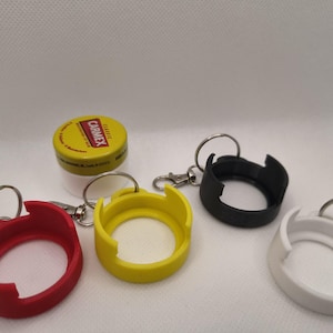 Könnte beinhalten: Eine Sammlung von Schlüsselanhängern in Rot, Gelb, Schwarz und Weiß, jeweils mit einem kreisförmigen Design und einem Metallschlüsselring. Ein gelb-weißes Carmex-Lippenbalsam-Behältnis ist ebenfalls vorhanden. Die Schlüsselanhänger scheinen 3D-gedruckt zu sein.