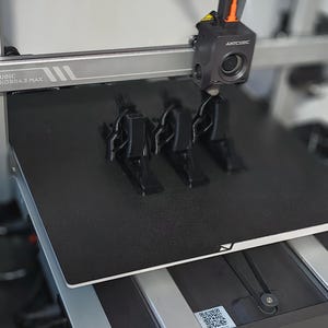 May include: A 3D printer, the Anycubic Kobra 3 Max, is shown printing three black plastic objects. The printer head is positioned above the objects, with the printer bed in the foreground. The printer is in operation.