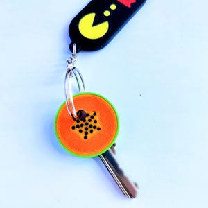 May include: A keychain featuring a black Pac-Man design with a yellow Pac-Man and a red ghost. The key has an orange and green papaya slice design. The keychain is attached to a silver key ring.