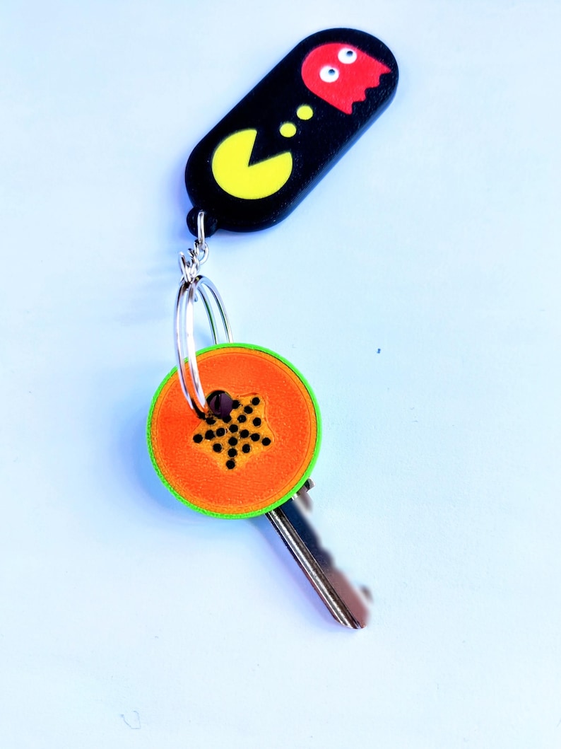 May include: A keychain featuring a black Pac-Man design with a yellow Pac-Man and a red ghost. The key has an orange and green papaya slice design. The keychain is attached to a silver key ring.