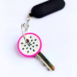 Puede incluir: Una llave con un llavero decorativo. El llavero es redondo con un borde rosa y un centro blanco con detalles negros en forma de semillas. La llave tiene una cadena plateada y un llavero rectangular negro.