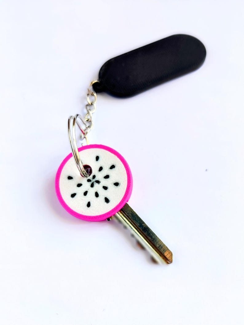May include: A key with a decorative charm. The charm is round with a pink border and a white center with black seed-like details. The key has a silver-colored chain and a black, rectangular fob.