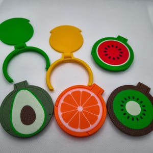 May include: A selection of fruit-shaped bottle stoppers. The stoppers are designed as a watermelon, orange, kiwi, and avocado. Each stopper has a hinged lid and a ring for attaching to a bottle. The colours include green, red, yellow, and brown.