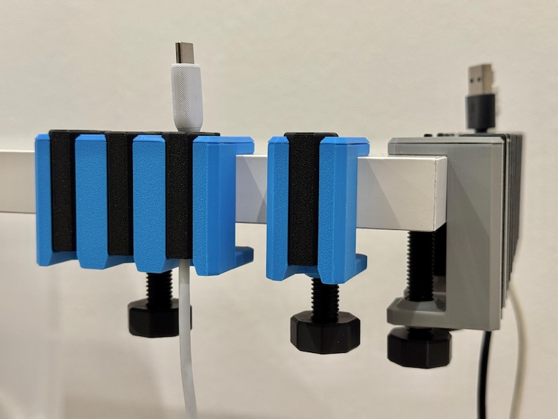 K&ouml;nnte beinhalten: Blaue und schwarze Kabelorganisatoren, an einer wei&szlig;en Stange befestigt. Die Organizer haben einen Schraubmechanismus. Ein wei&szlig;es USB-C-Kabel und ein schwarzes USB-Kabel sind eingesteckt.