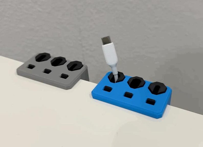 K&ouml;nnte beinhalten: Zwei Kabelorganisatoren, einer grau und einer blau, liegen auf einer wei&szlig;en Oberfl&auml;che. Der blaue Organizer h&auml;lt ein wei&szlig;es USB-C-Kabel. Jeder Organizer hat drei kreisf&ouml;rmige Schlitze und mehrere kleinere rechteckige Schlitze.