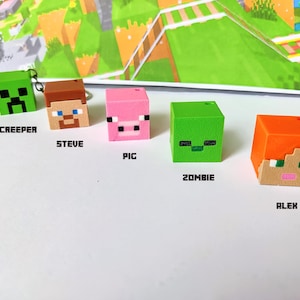 May include: Five Minecraft character cube figures: Creeper, Steve, Pig, Zombie, and Alex. Each figure is a different color, with pixelated facial features. The figures are arranged on a white surface with a colorful background.