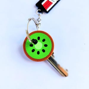 May include: A keychain featuring a kiwi fruit slice charm. The charm is green with black seeds and a brown rind. The keychain also includes a key and a black and red Top Dad tag.