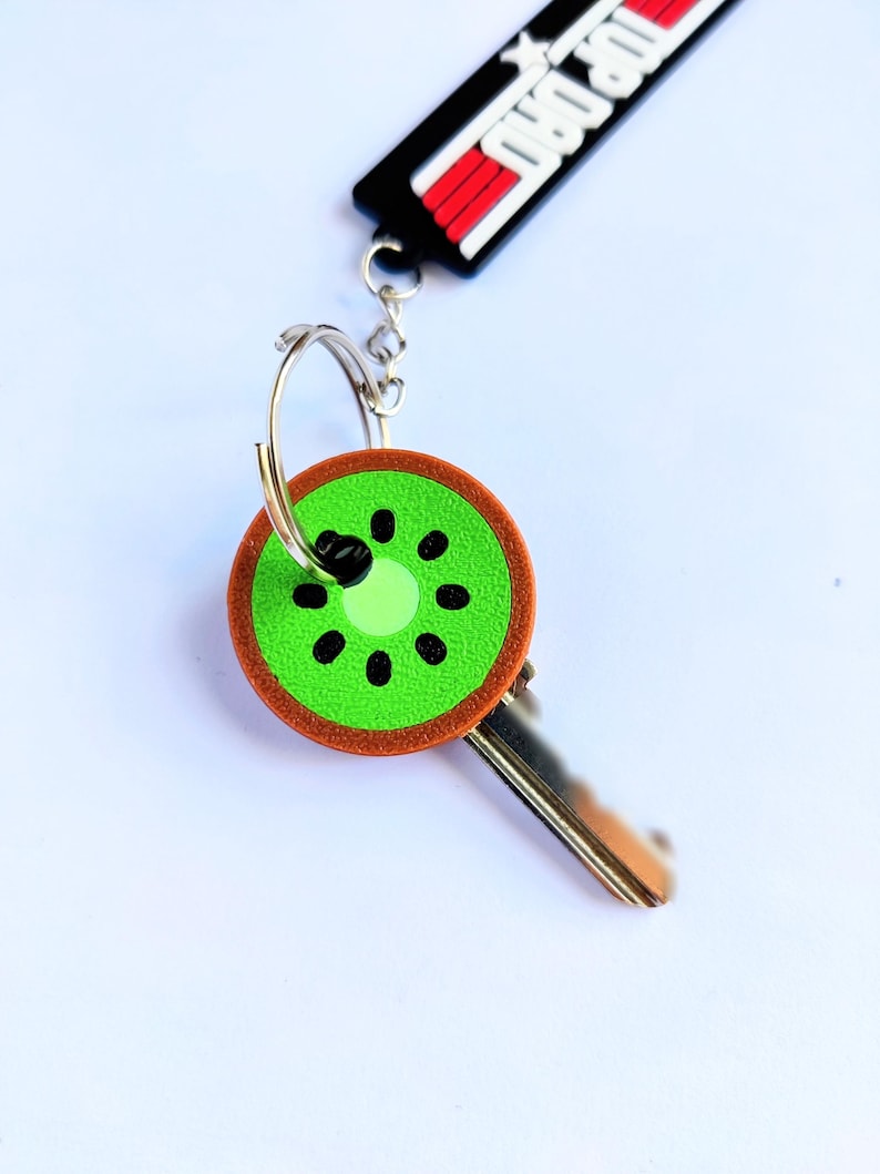 May include: A keychain featuring a kiwi fruit slice charm. The charm is green with black seeds and a brown rind. The keychain also includes a key and a black and red Top Dad tag.