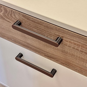 May include: Close-up of a cabinet with two drawers. The top drawer is made of wood-grain laminate, and the bottom drawer is white. Each drawer has a dark brown, rectangular handle. The cabinet top is a light cream color.