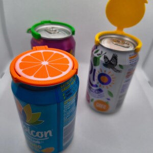 May include: Three beverage cans with colourful lids. One can has an orange slice lid, another has a green lid, and the third has a yellow lid. The cans are various colours, including blue, purple, and white.