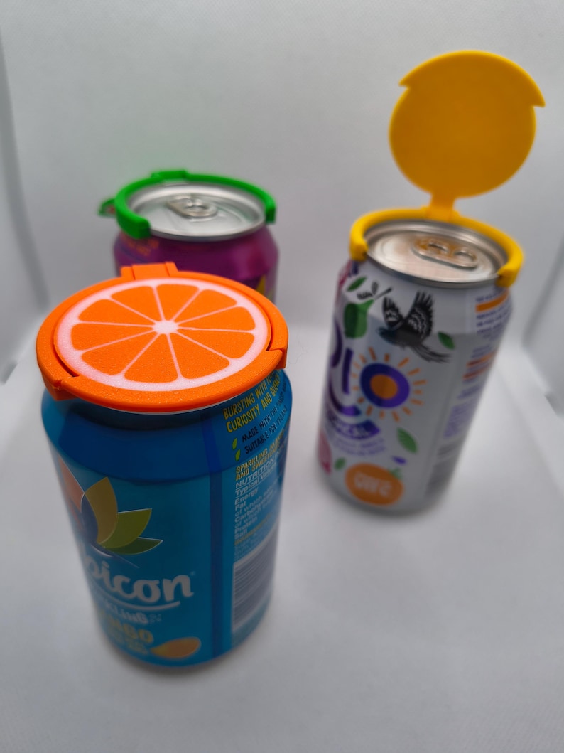 May include: Three beverage cans with colourful lids. One can has an orange slice lid, another has a green lid, and the third has a yellow lid. The cans are various colours, including blue, purple, and white.