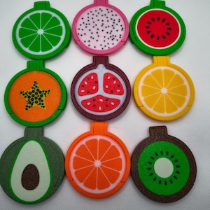 May include: Nine colourful fruit-shaped magnets are displayed. The magnets depict lime, dragon fruit, watermelon, papaya, pomegranate, lemon, avocado, orange, and kiwi. Each magnet is circular with a small rectangular tab at the top. Measurements are not applicable.