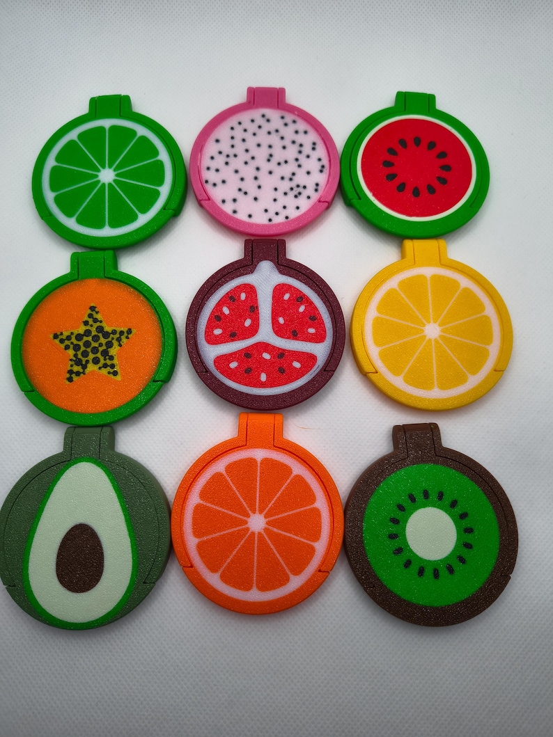 May include: Nine colourful fruit-shaped magnets are displayed. The magnets depict lime, dragon fruit, watermelon, papaya, pomegranate, lemon, avocado, orange, and kiwi. Each magnet is circular with a small rectangular tab at the top. Measurements are not applicable.