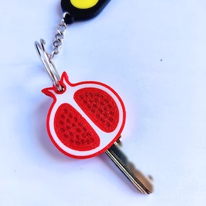 May include: A keychain featuring a red and white pomegranate charm and a black charm with a yellow Pac-Man design. The key is silver and the chain is also silver.