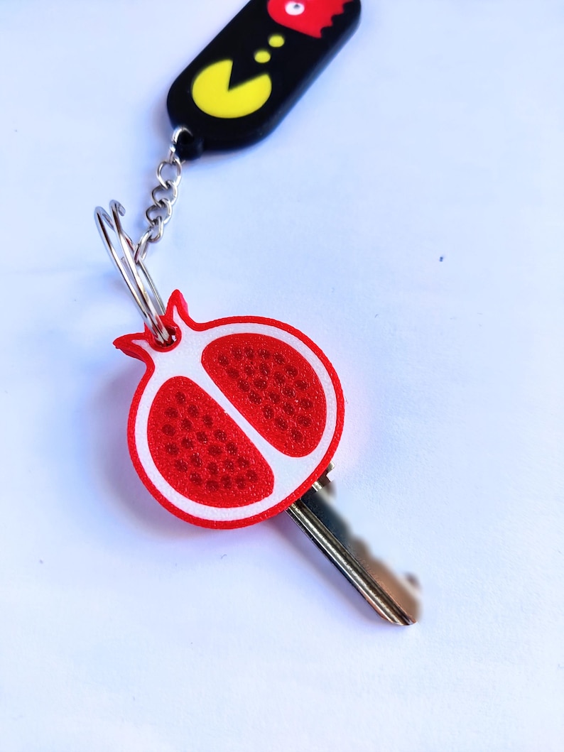 May include: A keychain featuring a red and white pomegranate charm and a black charm with a yellow Pac-Man design. The key is silver and the chain is also silver.