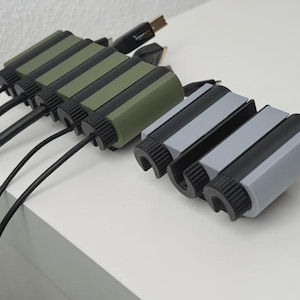 Puede incluir: Organizadores de cables en verde oliva y gris con detalles en negro. Los organizadores cilíndricos tienen ranuras para cables y están conectados a cables negros. Los artículos están sobre una superficie blanca.