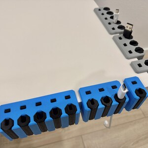 K&ouml;nnte beinhalten: Blaue und graue 3D-gedruckte Kabelorganisatoren mit mehreren Schlitzen f&uuml;r verschiedene Kabeltypen. Die Organizer sind f&uuml;r die Platzierung am Schreibtischrand konzipiert und halten Kabel ordentlich und zug&auml;nglich. Schwarze Akzente.