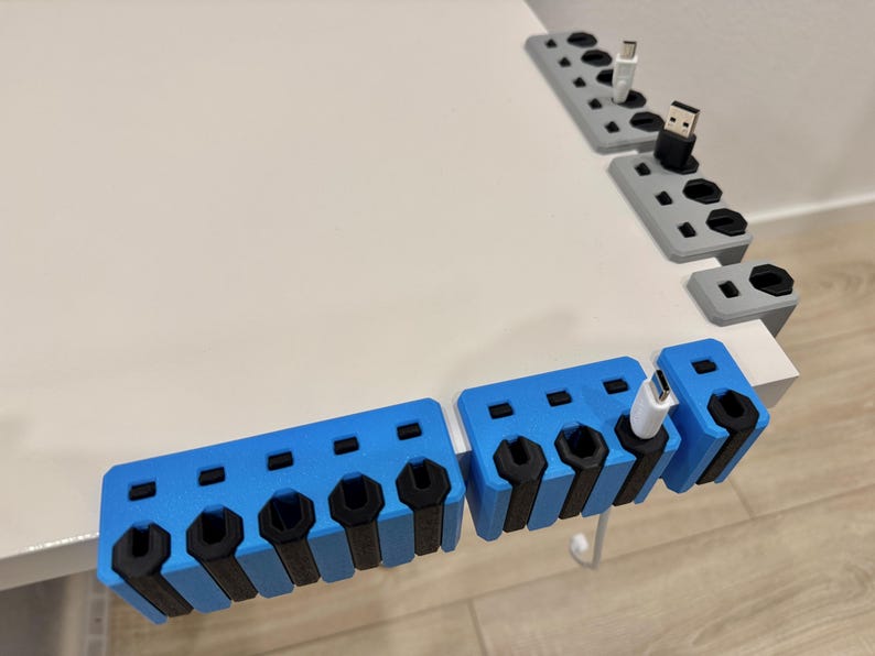 K&ouml;nnte beinhalten: Blaue und graue 3D-gedruckte Kabelorganisatoren mit mehreren Schlitzen f&uuml;r verschiedene Kabeltypen. Die Organizer sind f&uuml;r die Platzierung am Schreibtischrand konzipiert und halten Kabel ordentlich und zug&auml;nglich. Schwarze Akzente.