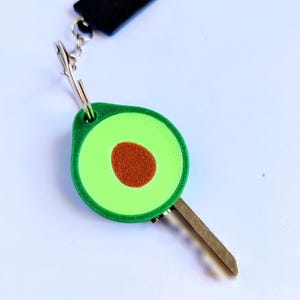 May include: A key with a green avocado-shaped key fob. The avocado has a brown seed in the center and a green border. The key fob is attached to a silver clasp and a black strap.