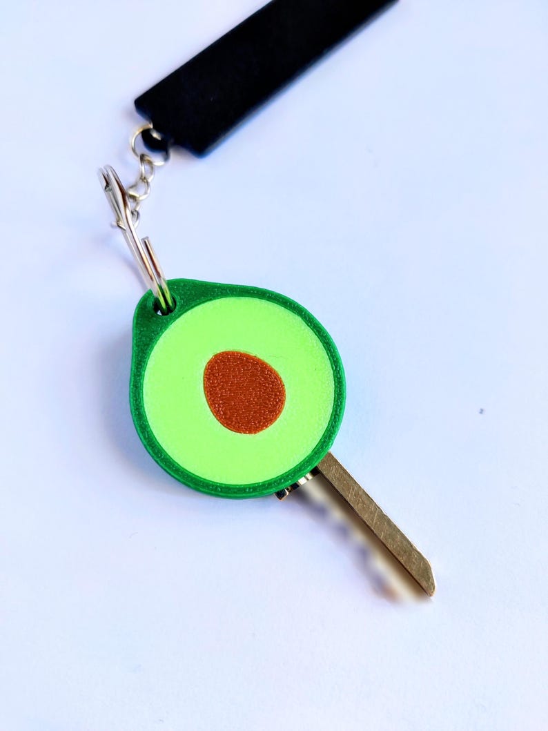 May include: A key with a green avocado-shaped key fob. The avocado has a brown seed in the center and a green border. The key fob is attached to a silver clasp and a black strap.