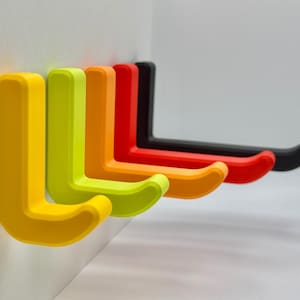 May include: Five L-shaped wall hooks in a row, in a gradient of colours: yellow, lime green, orange, red, and black. The hooks are mounted on a white wall. Each hook is designed for hanging items.
