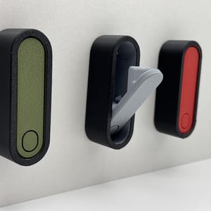 Op de afbeelding: Drie aan de muur gemonteerde haken met een modern design. De haken hebben een zwart frame met gekleurde inzetstukken: olijfgroen, grijs en rood. Elke haak heeft een cirkelvormig detail aan de onderkant. Eén haak is open en onthult een grijze haak.