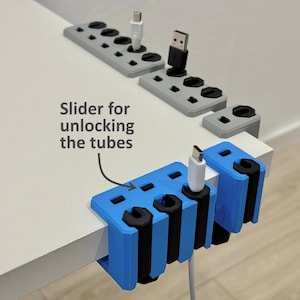 May include: A selection of blue and grey 3D-printed cable organisers. The blue organisers clamp onto a surface and hold cables. The image also shows grey organisers with multiple holes. Text on the image reads "Slider for unlocking the tubes."