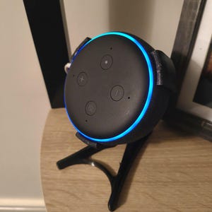 May include: A black smart speaker with a blue light ring, resting on a black stand. The speaker has control buttons on top. The stand has three legs and is on a light-colored wooden surface.