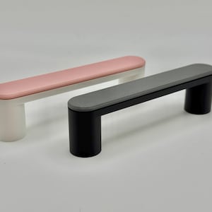 May include: Two cabinet pulls. One has a white base with a pink top, the other has a black base with a grey top. Both have a rectangular design with rounded edges and cylindrical supports.