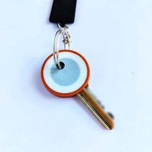 May include: A key with a decorative, round head featuring a light blue and white design, framed by a brown border. The key is attached to a silver keyring and a black lanyard.