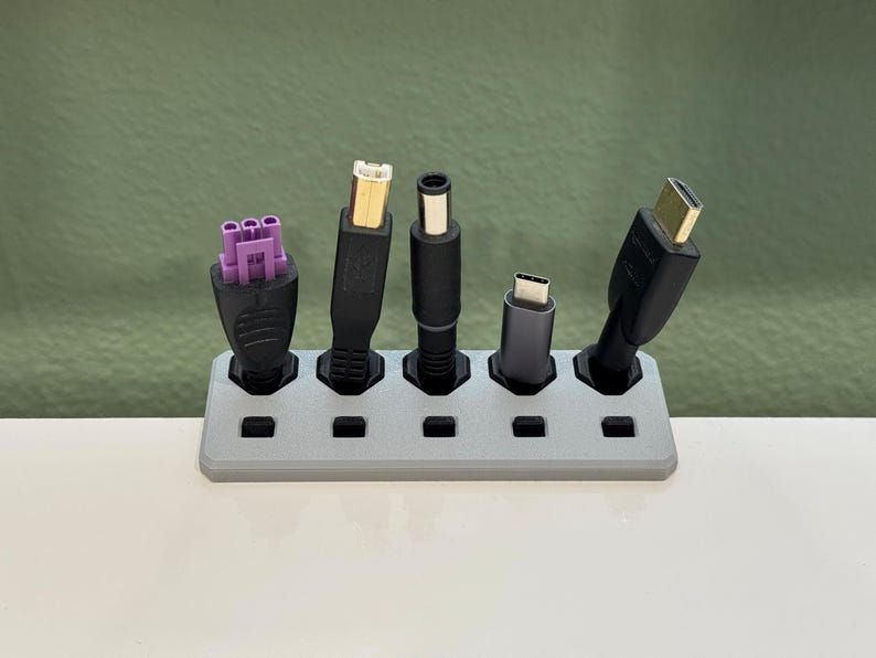 K&ouml;nnte beinhalten: Ein grauer, 3D-gedruckter Kabelorganizer, der verschiedene schwarze Kabel mit unterschiedlichen Steckertypen, einschlie&szlig;lich USB, und einen lila Stecker h&auml;lt. Der Organizer steht auf einer wei&szlig;en Oberfl&auml;che mit gr&uuml;nem Hintergrund.