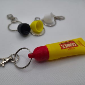Könnte beinhalten: Ein gelb-rotes Carmex Classic Lippenbalsam-Tuben-Schlüsselanhänger mit einem silbernen Schlüsselring und Verschluss. Auf der Tube ist das Carmex-Logo aufgedruckt. Im Hintergrund sind mehrere andere Schlüsselanhänger-Kappen zu sehen.