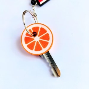 May include: A key with an orange slice design. The key has a silver metal blade and a round orange and white charm with a slice of orange design. The keychain also has a black and red tag that says "ROAD DAD".