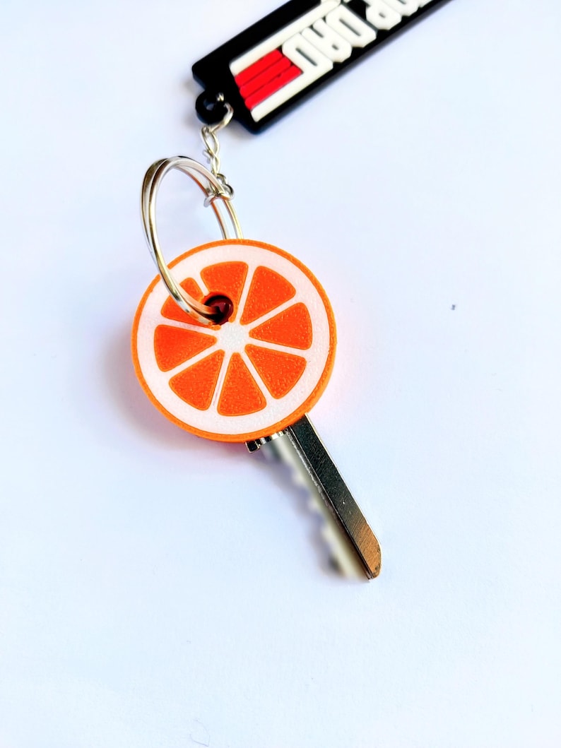 May include: A key with an orange slice design. The key has a silver metal blade and a round orange and white charm with a slice of orange design. The keychain also has a black and red tag that says "ROAD DAD".