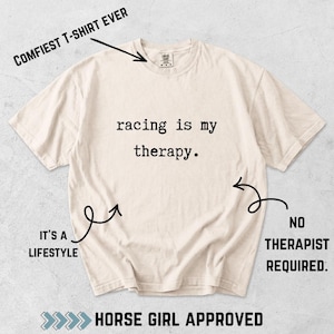 Op de afbeelding: Een crèmekleurig T-shirt met korte mouwen en de zwarte tekst 'racing is my therapy.' op de borst. De afbeelding bevat extra tekstannotaties: 'COMFIEST T-SHIRT EVER', 'IT'S A LIFESTYLE', 'NO THERAPIST REQUIRED.' en 'HORSE GIRL APPROVED'.