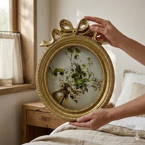 Op de afbeelding: Een goudkleurige, ronde fotolijst met een strik aan de bovenkant. De lijst bevat een bloemenprint met witte bloemen en groene bladeren in een vaas. De lijst wordt door twee handen vastgehouden.