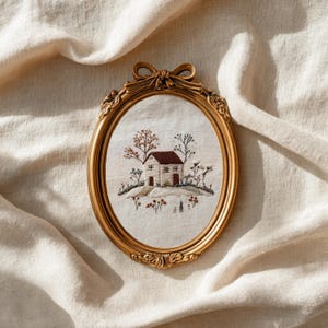 May include: An oval, gold-colored framed embroidery featuring a quaint house with a brown roof, surrounded by embroidered trees and flowers. The frame has a decorative bow at the top. The artwork is set against a cream-colored fabric.
