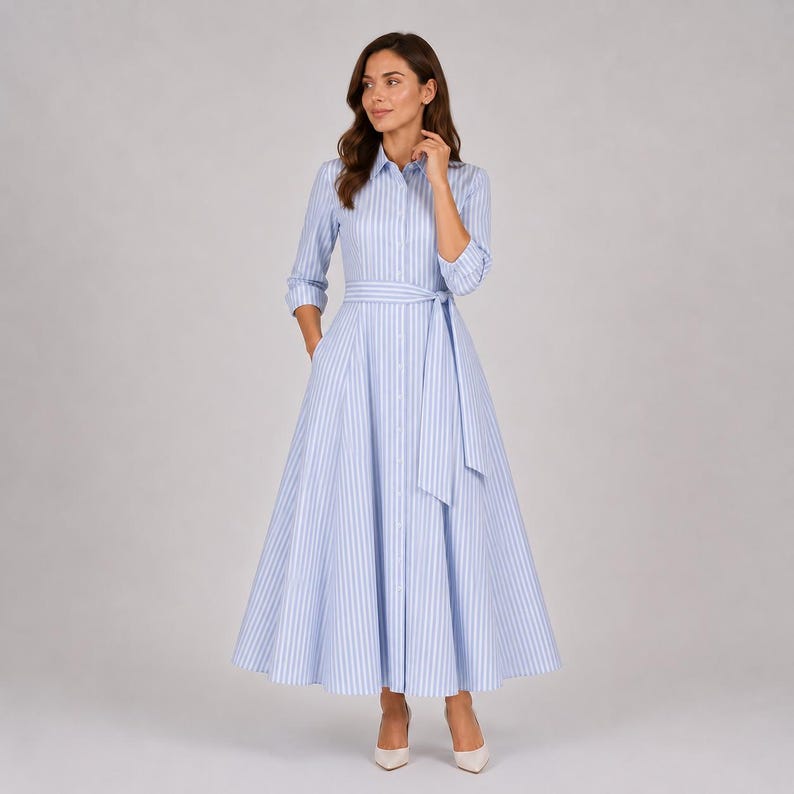May include: A long-sleeved shirt dress featuring a light blue and white vertical striped pattern. The dress has a collared neckline, a button-down front, and a matching fabric tie belt at the waist, with a full, flared skirt that reaches the ankles.