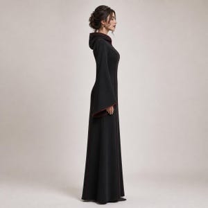 May include: A floor-length black dress featuring a hood, long flared sleeves, and a fitted bodice. The garment is accented with thin dark red piping along the seams, the hood edge, and the sleeve cuffs, creating a structured and clean silhouette.