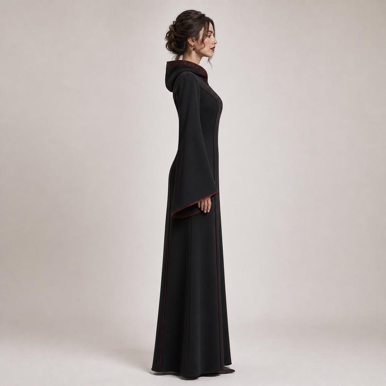 May include: A floor-length black dress featuring a hood, long flared sleeves, and a fitted bodice. The garment is accented with thin dark red piping along the seams, the hood edge, and the sleeve cuffs, creating a structured and clean silhouette.