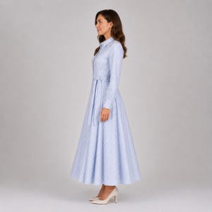 May include: A long-sleeved, ankle-length shirt dress featuring a vertical light blue and white striped pattern. The dress has a classic collar, a button-down front, and a matching fabric tie belt at the waist, creating a full, flared skirt silhouette.