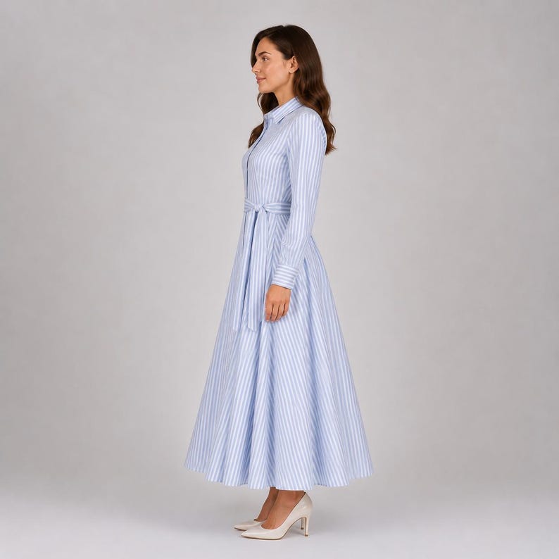 May include: A long-sleeved, ankle-length shirt dress featuring a vertical light blue and white striped pattern. The dress has a classic collar, a button-down front, and a matching fabric tie belt at the waist, creating a full, flared skirt silhouette.