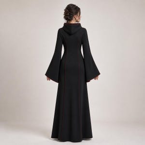 May include: A floor-length black dress featuring a hood, long flared bell sleeves, and thin dark red piping along the seams. The garment has a fitted bodice that transitions into a flared skirt, showcasing a solid black fabric throughout.