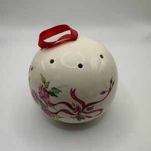 May include: A spherical cream-coloured ceramic pomander featuring a hand-painted floral design with pink roses, green leaves, and magenta ribbons. The object has several small circular ventilation holes and a red fabric ribbon loop for hanging.