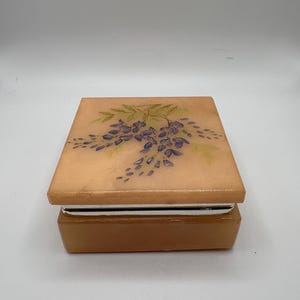 May include: A square, light orange stone box with a smooth, polished finish. The lid features a painted illustration of purple wisteria flowers with green leaves. A thin, reflective silver-toned metal band separates the lid from the base.