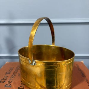 May include: An oval-shaped brass basket featuring a polished metallic finish and a single arched handle attached to the sides with small rivets. The container has a smooth, rounded rim and a solid base, showcasing a warm, reflective golden tone.