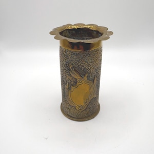 May include: A cylindrical brass vase featuring a textured, hammered finish on the body and a smooth, scalloped rim. The center of the body displays a polished, shield-shaped emblem with decorative scrollwork accents on the sides.